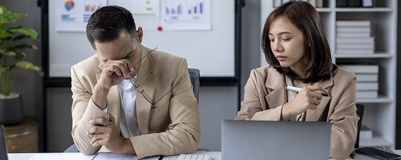 Two people in a business meeting, one of them is crying Two people in a business meeting, one of them is crying. The man is wearing a suit and the woman is wearing a brown jacket. They are sitting at a desk with a laptop and a stack of papers