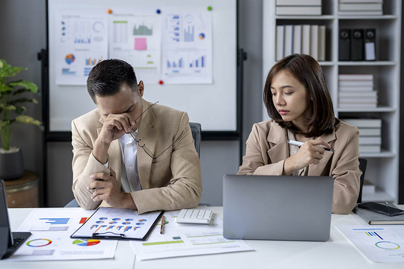 Two people in a business meeting, one of them is crying. The man is wearing a suit and the woman is wearing a brown jacket. They are sitting at a desk with a laptop and a stack of papers
